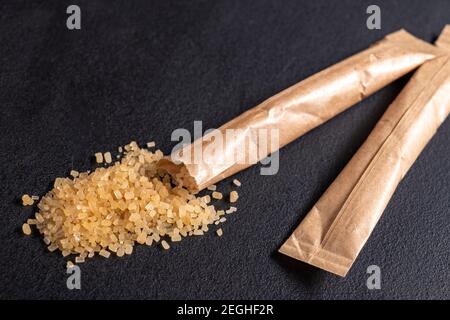 Spilled cane sugar from a small sachet. Preparation for sweetening beverages in a small package. Dark background. Stock Photo
