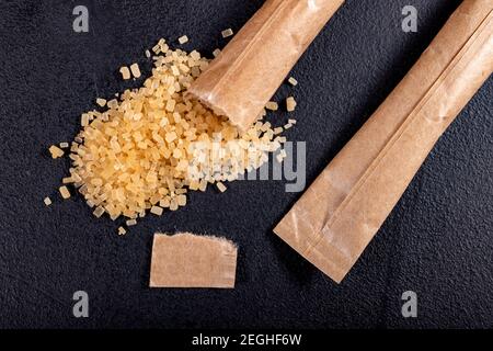 Spilled cane sugar from a small sachet. Preparation for sweetening beverages in a small package. Dark background. Stock Photo