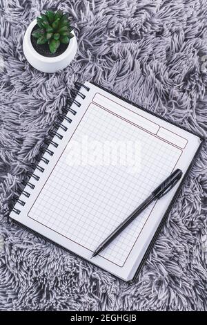 Aerial view of an open notebook with a pen on top and a green plant next to it, on a gray fuzzy surface Stock Photo