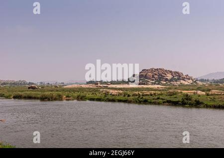 Anegundi, Karnataka, India - November 9, 2013: Gray Tungabhadra river. Wide landscape with Sooryanarayana Temple complex on its green island under lig Stock Photo