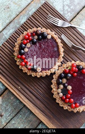 Small vegan tarts made of nuts and berry jam decorated with black, red and white currants on oak cutting board overhead view Stock Photo