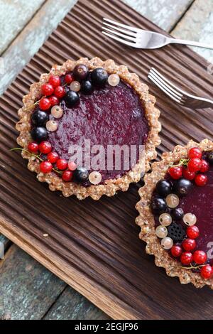 Small vegan tarts made of nuts and berry jam decorated with black, red and white currants on oak cutting board overhead view Stock Photo