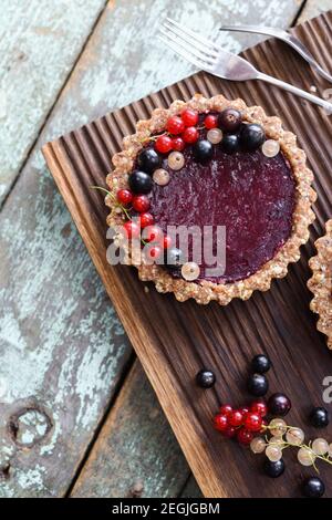 Small vegan tarts made of nuts and berry jam decorated with black, red and white currants on oak cutting board overhead view Stock Photo