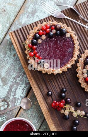 Small vegan tarts made of nuts and berry jam decorated with black, red and white currants on oak cutting board overhead view Stock Photo