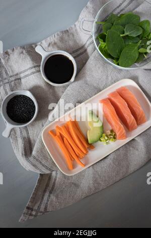 Overhead view of a tray full of salmon, avocado and carrots next to a spinach bowl and a soy cup and a black sesame cup on a grey cloth. Stock Photo