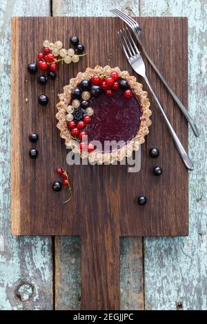 Small vegan tarts made of nuts and berry jam decorated with black, red and white currants on oak cutting board overhead view Stock Photo