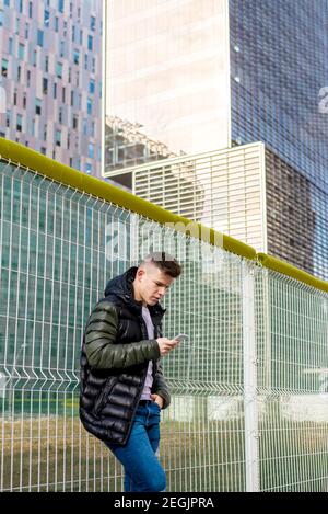 Young man leaning on metallic fence, using smartphone in a city. Stock Photo