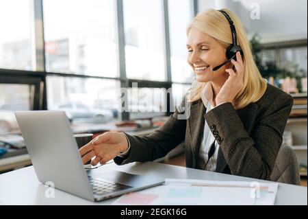 Elegant female employee of support line in headset is using laptop, sitting at desk. Middle aged woman manager conducts online business meeting, communication concept Stock Photo