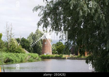 A traditional windmill (wind pump) at a bend in a tranquil river. Built on the riverbank, partially obscured by willow trees. Placid landscape Stock Photo