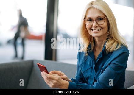 Smiling elegant middle aged caucasian woman in formal wear and eyeglasses uses smartphone, browsing internet or chatting with friends, looks at the camera, smiling Stock Photo