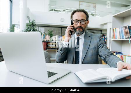 Concentrated entrepreneur is sitting at the desk in office, using cellphone. Senior successful man talks with colleague by smartphone, discussing new business plan, making notes Stock Photo