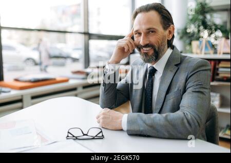 Portrait of successful happy entrepreneur at the workplace. Smiling middle aged businessman sits at the desk, looks at the camera, wearing stylish formal suit Stock Photo
