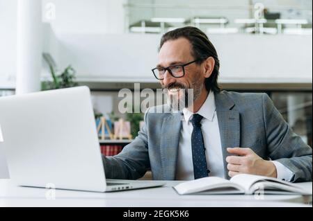 Satisfied businessman is sitting at the desk in office, happily looks at the laptop screen. Middle aged bearded entrepreneur is using computer, developing an app or project Stock Photo