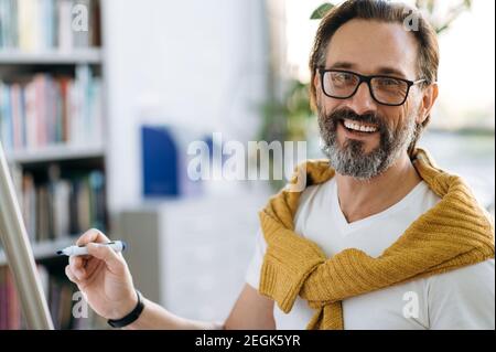 Portrait of a happy male employee or teacher in eyeglases, holds a marker in hand, writing on flipchart financial graphs or theme of lecture, looks and smiles at camera Stock Photo