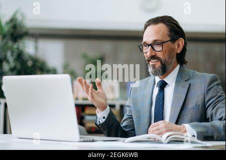 Satisfied middle aged entrepreneur at online business meeting, discussing with coworkers. Successful male employee in formal suit is sitting at the desk in office, having a video call, smiling Stock Photo