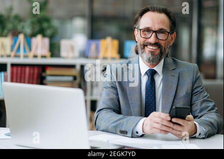 Smiling grey-haired business man looks directly at the camera, sits at the work desk. Happy influential male employee is using smartphone, and friendly smiles Stock Photo