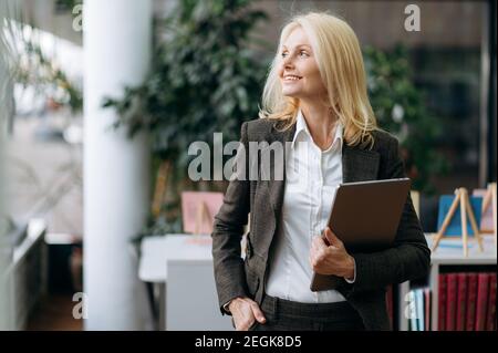 Elegant middle aged lady in stylish suit stands in modern office, smiling. Beautiful successful businesswoman is working as office employee, holding laptop in arms, looks away Stock Photo
