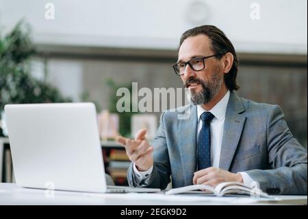Elegant senior businessman is having a conversation on video conference. Concentrated male ceo communicate with colleagues, discussing important project Stock Photo