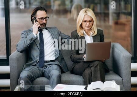 Serious business partners are sitting on the sofa in modern office, using laptop and smartphone. Mature colleagues working on corporate project, discussion ideas, partnership concept Stock Photo