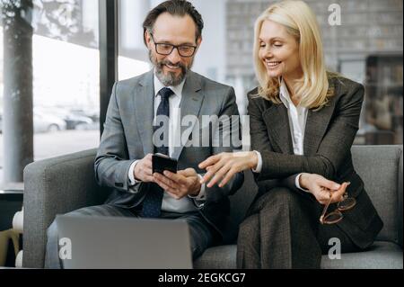 Happy friendly business partners using smartphone, browsing internet. Successful co-workers in formal suits sitting on the sofa, take a break from work, discussing information Stock Photo