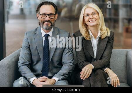 Portrait of two mature elegant business people in stylish suits at the modern office. Friendly successful colleagues sitting on the couch, looking directly at the camera, friendly smiling Stock Photo