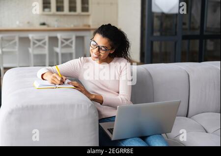 Positive smart young African American female freelancer in glasses sitting on the couch with laptop on the lap, studying from home and taking notes to Stock Photo