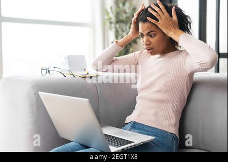 Shocked young African American female freelancer student with curly hair holding head in hands, sitting on the couch with laptop on lap, stressing out Stock Photo