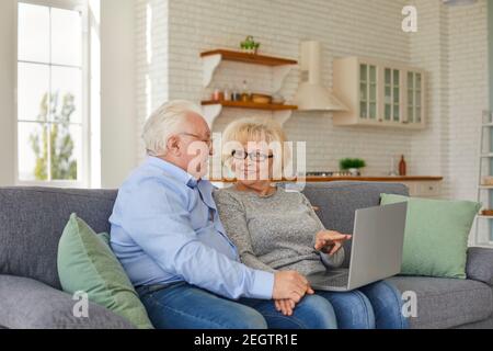 Smiling mature wife and husband looking at laptop screen sitting on sofa in cozy home. Stock Photo
