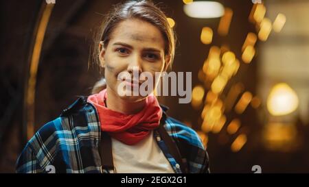 Young Beautiful Empowering Woman with Ear Piercing Gently Smiles at the Camera. Authentic Fabricator Wearing Work Clothes in a Metal Workshop. Sparks Stock Photo