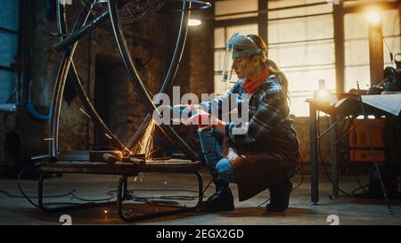 Young Contemporary Female Artist is Polishing a Metal Tube Sculpture with an Angle Grinder in a Workshop. Pretty Empowering Woman with Ear Piercing. Stock Photo