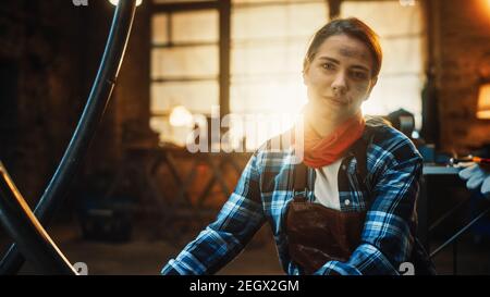 Young Beautiful Empowering Woman with Ear Piercing Gently Smiles at the Camera. Authentic Fabricator Wearing Work Clothes in a Metal Workshop. Stock Photo