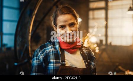 Young Beautiful Empowering Woman with Ear Piercing Gently Smiles at the Camera. Authentic Fabricator Wearing Work Clothes in a Metal Workshop. Sparks Stock Photo