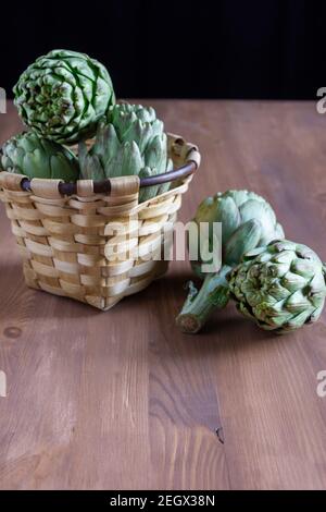 Top view of rustic basket with artichokes on wooden table with two artichokes, black background, vertical Stock Photo