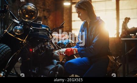 Young Beautiful Female Mechanic is Fixing a Custom Bobber Motorcycle. Talented Girl Wearing a Blue Jumpsuit. She Uses a Ratchet Spanner. Creative
