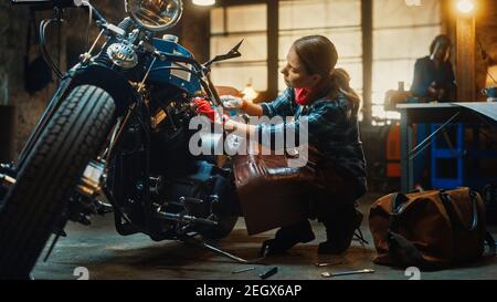 Young Beautiful Female Mechanic is Working on a Custom Bobber Motorcycle. Talented Girl Wearing a Checkered Shirt and an Apron. She Uses a Spanner to