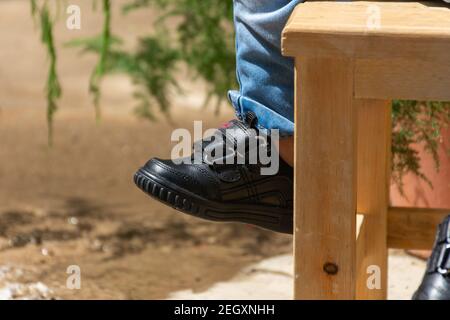 Baby feet of boy wears black leather shoes sitting on a wooden chair with copy space on the left Stock Photo