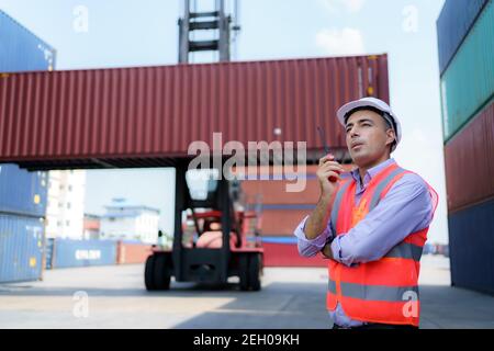 Caucasian foreman man is using walkie talkie to command his workers with crane lifting container in background in container depot terminal. Stock Photo