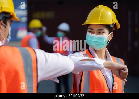 Asian worker new normal greeting by elbows bump wear safety helmet and mask in container depot terminal for prevent coronavirus and social distancing Stock Photo