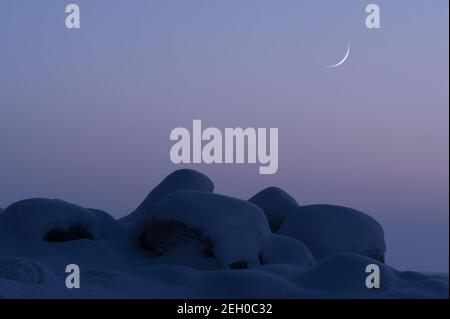 Winter landscape at twilight. Crescent new moon in the sky behind snow covered boulders. Stock Photo