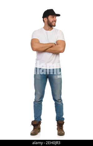 Casual young man in black cap, jeans and white t-shirt is standing with arms crossed, looking at the side and smiling. Full length studio shot isolate Stock Photo