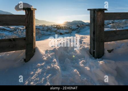 Snow covered rural winter landscape seen through fence during sunset with sun star between alpine mountains, Wildermieming, Tirol, Austria Stock Photo