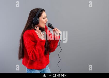 young, emotional brunette with headphones and a microphone dressed in a red sweater sings karaoke, isolated on gray Stock Photo