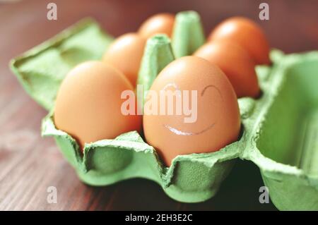 Group of brown chicken eggs in an green open form work on brown background. The closest egg is painted with white smiley face. Front view. Stock Photo