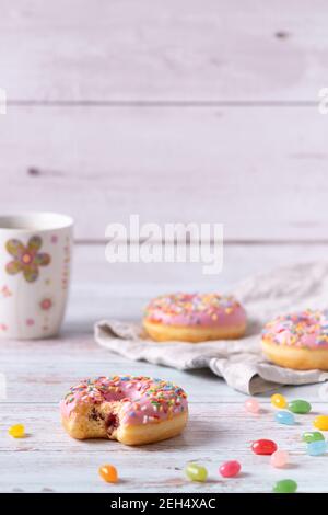 Bitten strawberry donuts with pink icing, colorful sprinkles and jelly beans on wooden background. Sweet pastry as a snack for children's birthday par Stock Photo