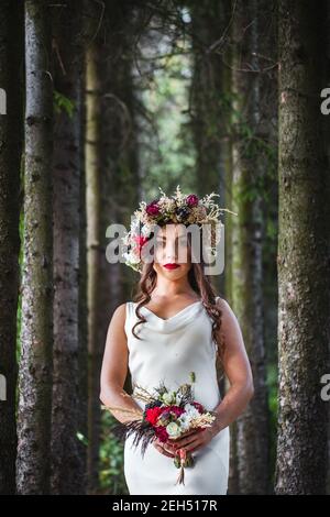 Wedding. Young beautiful bride in white dress and wreath standing at green lawn between trees. Summer full length atmospheric portrait portrait. Stock Photo