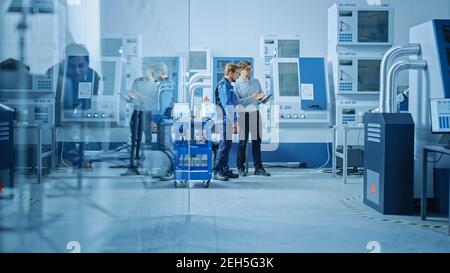 In Factory Workshop: Workers Use High-Tech Industrial CNC Machinery. Two Engineering Professional Talking and Using Digital Tablet. Stock Photo