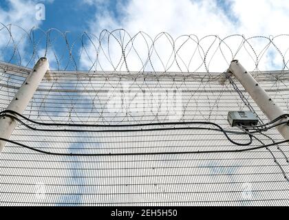 High security prison fence with razor barbed wire on top looking up from ground at solid metal alarmed mesh fencing cables and blue summer sky freedom Stock Photo