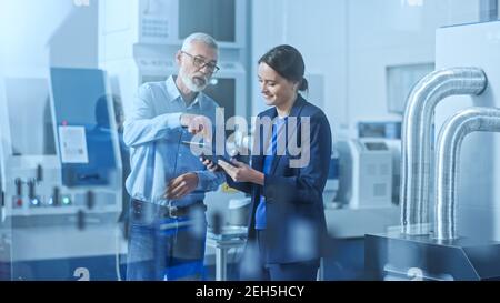 Modern Factory: Female Engineer, Male Project Manager Standing in High Tech Development Facility, Talking and Using Tablet Computer. Contemporary Stock Photo