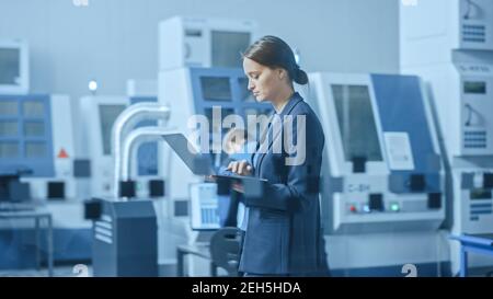 Modern Factory: Confident Female Engineer Walks Through Facility, Holding Laptop, Inspecting and Monitoring Workshop Workings. Manufactory Has Stock Photo