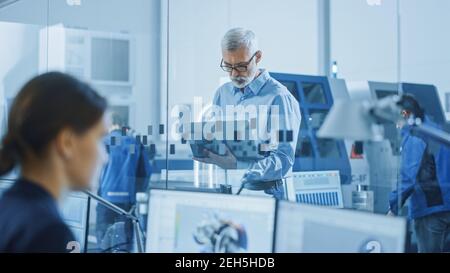 Modern Factory: Senior Engineer Looks at Camera, Holding Laptop, Inspecting, Overlooking Workshop Operation. Manufactory Has Professionals Working on Stock Photo